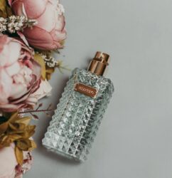 A luxurious glass perfume bottle beside delicate pink peonies on a gray background, showcasing elegance and fragrance.