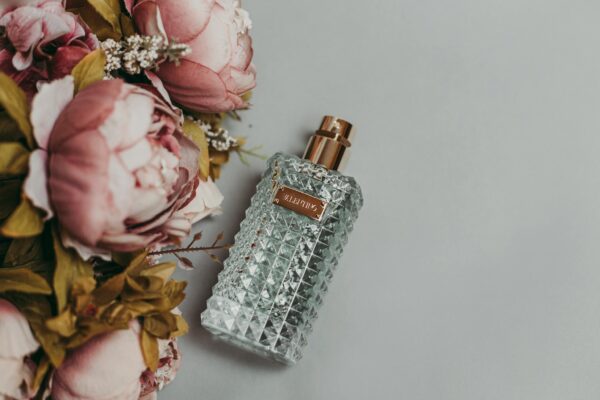 A luxurious glass perfume bottle beside delicate pink peonies on a gray background, showcasing elegance and fragrance.